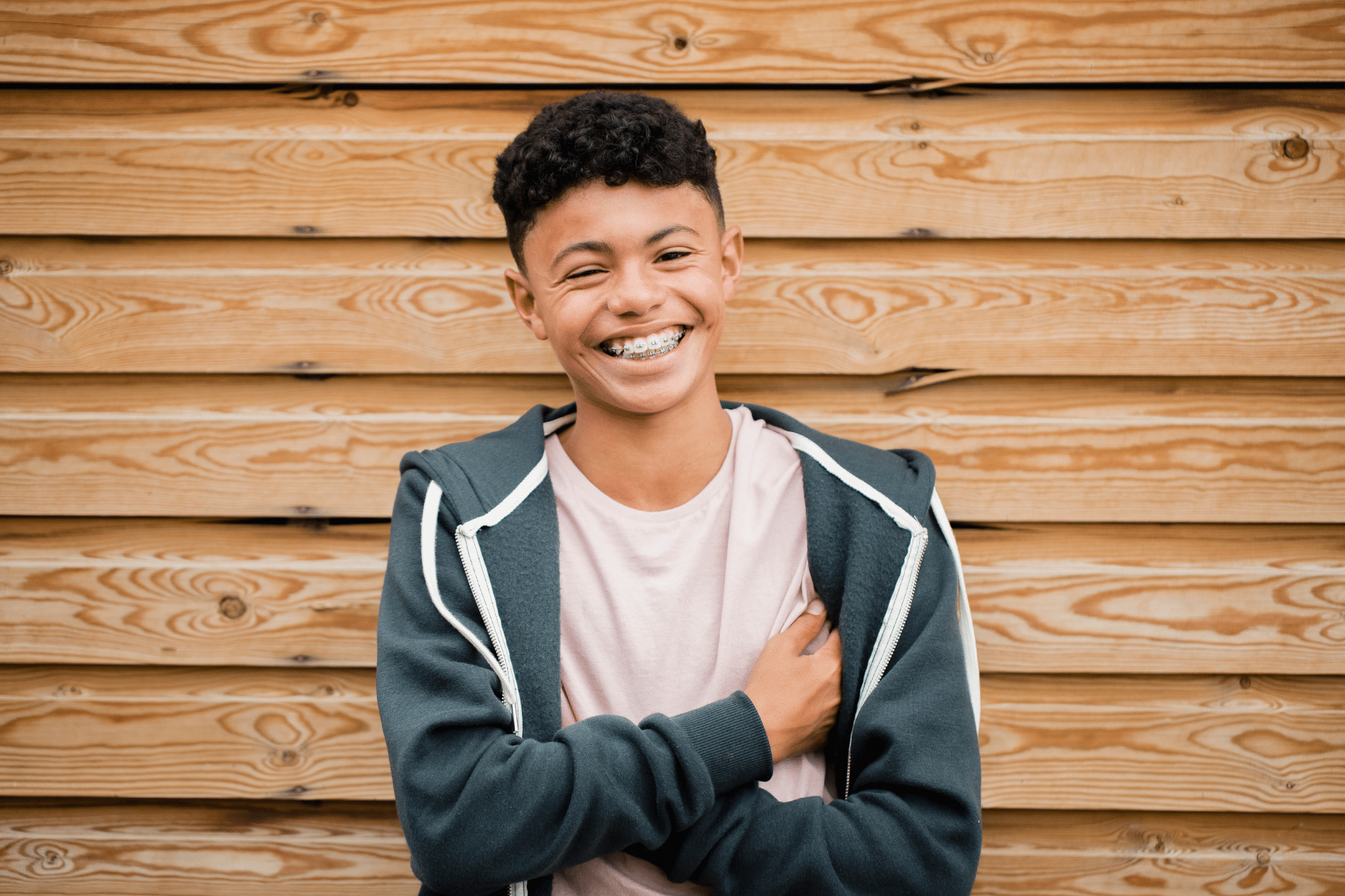 Una adolescente sonriente con aparato dental y los brazos cruzados delante de una pared de madera